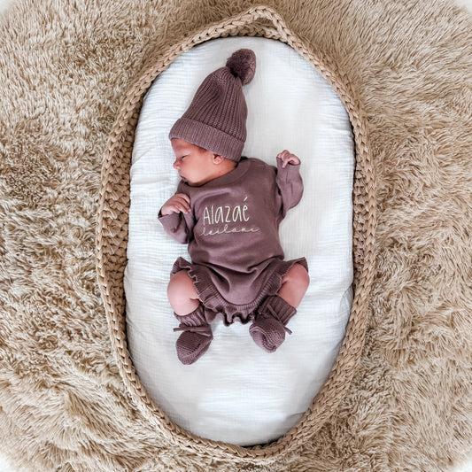 Newborn baby in a embroidered outfit and hat lying on a white blanket with a textured brown background
