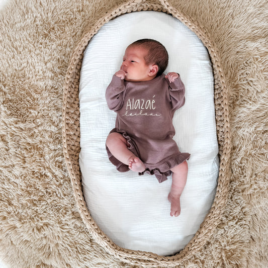 Newborn baby in an embroidered outfit lying on a white cushion inside a woven basket.