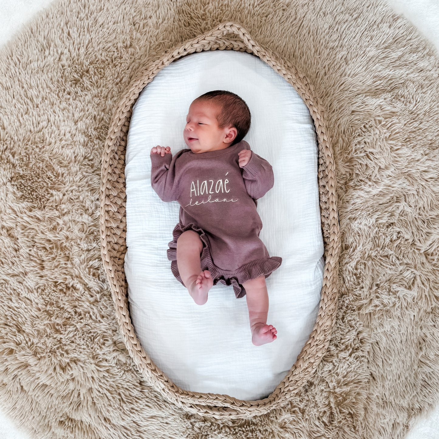 Newborn baby in a purple outfit with 'Alazae' name, lying on a white cushion surrounded by beige fabric.
