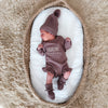 Newborn baby in a embroidered name outfit and hat lying on a white cushion with a textured brown background.