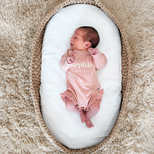 Newborn baby in a pink embroidered onesie lying on a white cushion inside a woven basket on a fluffy beige surface.
